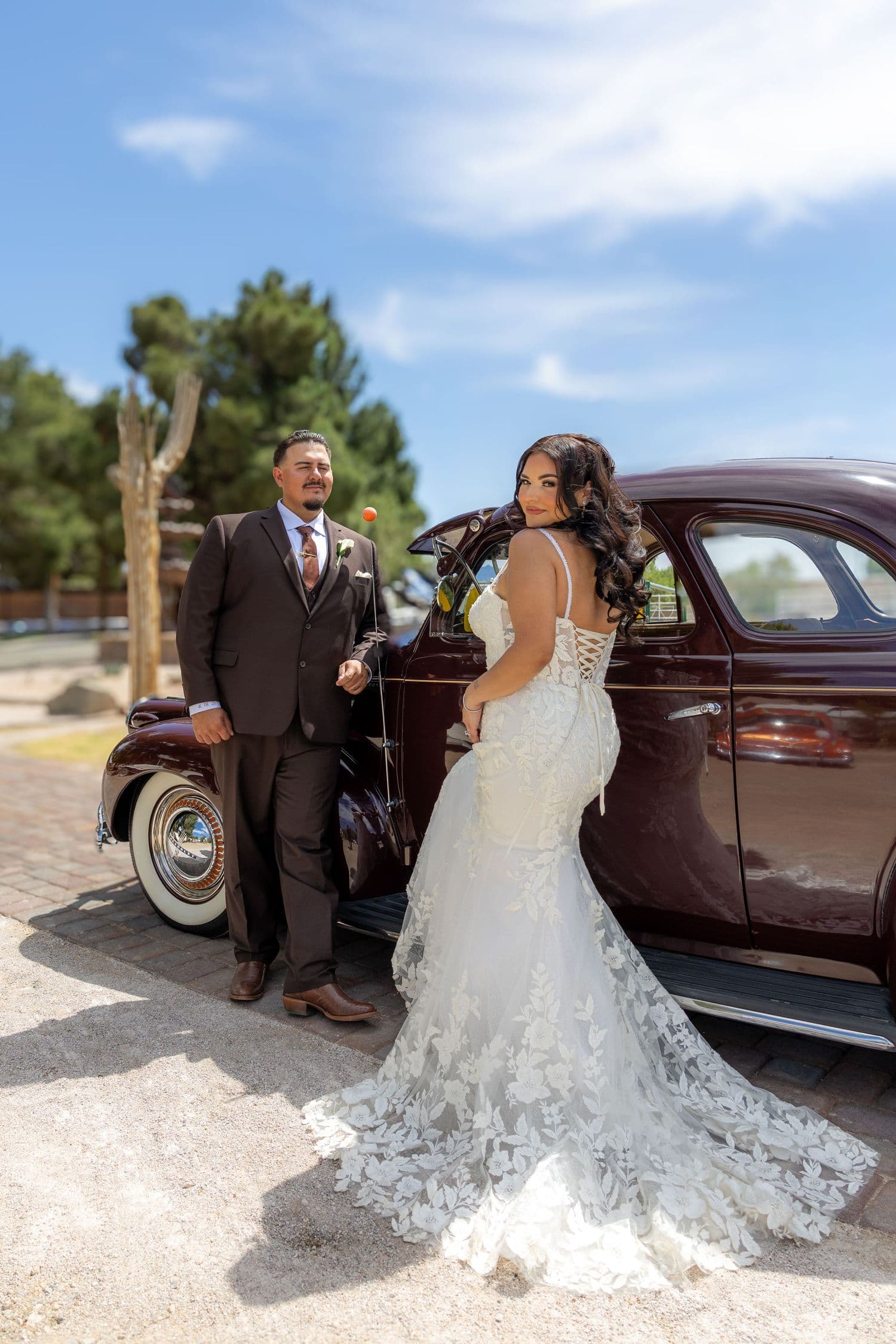 Bride in a lace mermaid gown and groom in a brown suit beside a vintage maroon car